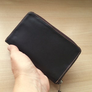 May include: A rectangular black leather wallet featuring a dark brown zipper closure along three sides. The wallet is constructed with visible brown stitching along the edges, providing a contrast to the smooth, dark surface of the leather material.