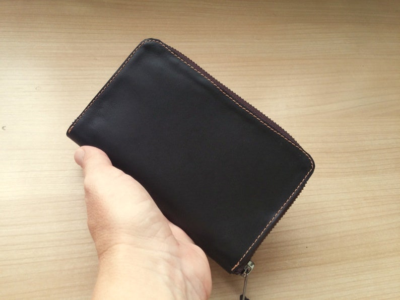 May include: A rectangular black leather wallet featuring a dark brown zipper closure along three sides. The wallet is constructed with visible brown stitching along the edges, providing a contrast to the smooth, dark surface of the leather material.