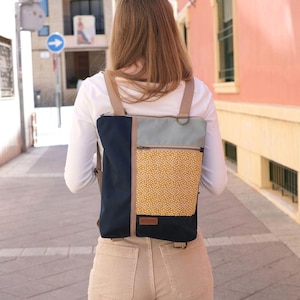 Puede incluir: Una mochila multicolor que presenta un panel lateral azul marino oscuro, una sección superior gris claro y un bolsillo inferior amarillo mostaza con un patrón de puntos blancos. Incluye correas beige, una cremallera superior y una etiqueta marrón.