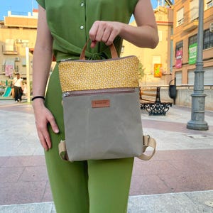 May include: A rectangular backpack featuring a grey lower section and a mustard yellow upper section with a white geometric pattern. The bag includes a horizontal zippered pocket, a brown leather brand tag, and tan adjustable shoulder straps.