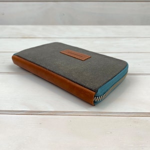 May include: A rectangular wallet featuring a dark gray canvas body, brown leather trim along the side, and a light blue zipper closure. A small brown leather rectangular patch is centered on the top surface with the text handmade embossed on it.