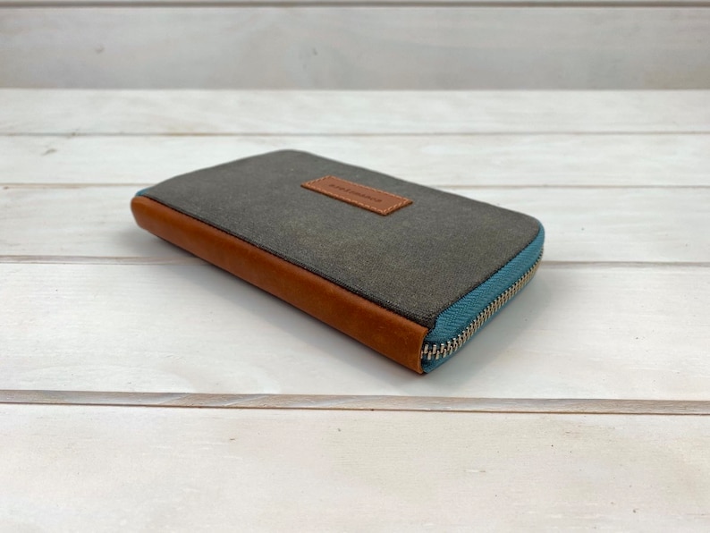 May include: A rectangular wallet featuring a dark gray canvas body, brown leather trim along the side, and a light blue zipper closure. A small brown leather rectangular patch is centered on the top surface with the text handmade embossed on it.
