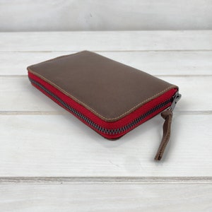 May include: A rectangular brown leather wallet featuring a vibrant red fabric zipper border and a dark metal zipper pull with a matching brown leather tab. The wallet has visible stitching along the edges and a smooth, matte finish on the surface.