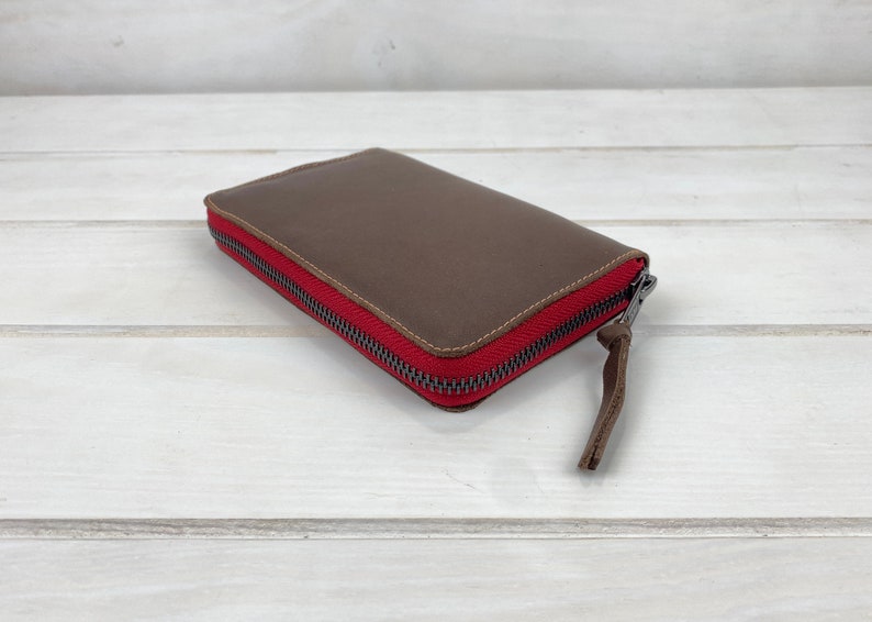 May include: A rectangular brown leather wallet featuring a vibrant red fabric zipper border and a dark metal zipper pull with a matching brown leather tab. The wallet has visible stitching along the edges and a smooth, matte finish on the surface.