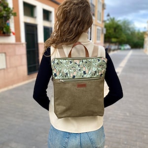 Puede incluir: Una mochila de forma cuadrada con una sección inferior de color verde oliva sólido y un panel superior con un patrón de hojas botánicas en verde y rosa. La bolsa incluye un bolsillo horizontal con cremallera, una etiqueta de marca de cuero marrón.