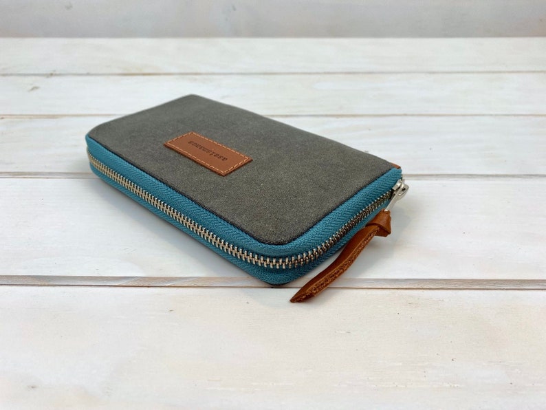 May include: A rectangular wallet featuring a dark gray top panel and a light blue side panel with a silver metal zipper. A brown leather rectangular patch is stitched onto the top surface, and a brown leather pull tab is attached to the zipper.
