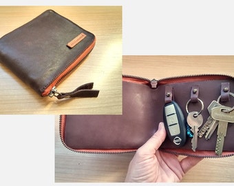 May include: A dark brown leather key case with a zipper closure and orange interior lining. The exterior features a small leather tag, while the interior contains three metal key rings and a leather strap for securing keys inside the case.