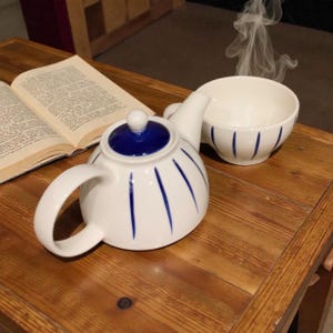 Op de afbeelding: Een witte keramische theepot en kop en schotel set met blauwe accenten. De theepot heeft een blauw deksel en verticale blauwe strepen. De theekop heeft vergelijkbare blauwe strepen. Stoom komt uit de theekop, wat duidt op een warme drank. Een open boek ligt ernaast.