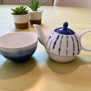 May include: A white ceramic teapot and teacup set with blue detailing. The teapot features a blue lid and vertical blue stripes. The teacup has a blue ombre design. Two small potted plants are in the background.