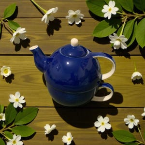 Puede incluir: Una tetera de cerámica azul cobalto con asa y boquilla blancas, sobre una superficie de madera. Flores blancas y hojas verdes rodean la tetera, creando una escena natural y acogedora.