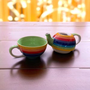 Op de afbeelding: Een keramische theekop en theepot set. De theekop heeft een groene binnenkant en een regenboog gestreepte buitenkant. De theepot is rond met een regenboog gestreept ontwerp en een groen handvat en tuit.