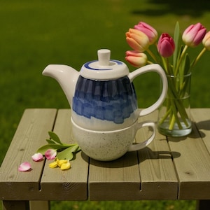May include: A white ceramic teapot with a blue watercolor design sits atop a matching speckled teacup. The teapot has a curved spout and handle. A vase of pink and yellow tulips and flower petals are on a wooden table.