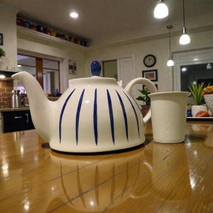 Op de afbeelding: Witte keramische theepot met blauwe verticale strepen en een bijpassende mok. De theepot heeft een gebogen tuit en een blauwe knop op het deksel. De mok is effen wit. Beide staan op een houten oppervlak.