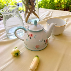 Puede incluir: Una tetera de cerámica blanca con asa y borde azules, decorada con rosas rosas y hojas verdes. Una taza a juego con borde dorado está cerca. También hay un tarro de cristal transparente y un jarrón con tallos secos.