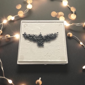 May include: A square, white coaster with a textured surface. The coaster features a dark gray, abstract design resembling a bird in flight. The coaster is set against a dark background, with string lights in the background.