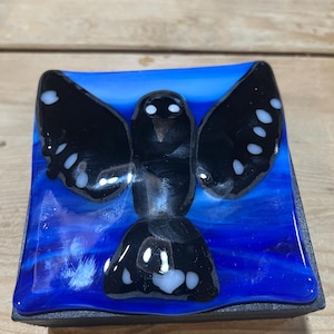 May include: A square fused glass piece featuring a black bird with white spots on its wings and tail, set against a vibrant blue background with swirling light and dark blue tones. The bird is centered, with wings spread wide in a flying pose.