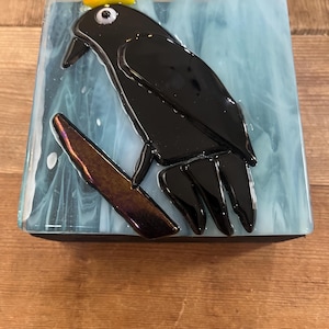 May include: A square fused glass art piece featuring a black bird perched on a brown branch against a light blue and white swirled background. The bird wears a small yellow, red, and green crown on its head and has a single white eye detail.