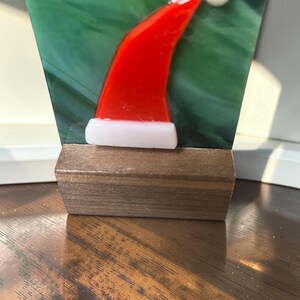 May include: A fused glass art piece featuring a red and white Santa hat design set against a green swirled background. The glass panel is mounted into a rectangular dark brown wooden base, creating a stable display for this holiday-themed object.