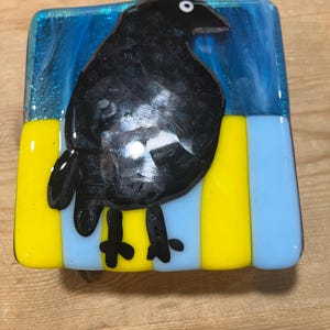 May include: A square fused glass art piece featuring a black bird with a white eye. The bird is positioned against a dark blue background, standing on a base of alternating vertical yellow and light blue stripes. The glass has a smooth finish.