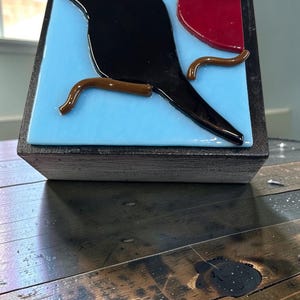 May include: A square decorative block featuring a fused glass panel with a light blue background. A black bird silhouette is perched on a brown branch, with a solid red circular shape positioned in the upper right corner of the glass surface.