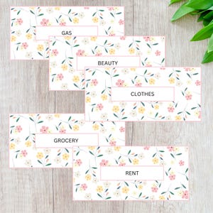 May include: Floral patterned budget envelopes with labels for "GAS", "BEAUTY", "CLOTHES", "GROCERY", and "RENT". The envelopes have a white background with pink, yellow, and white flowers and green leaves. Each label is in a white rectangle with a pink border.