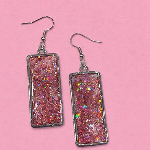 May include: A pair of rectangular dangle earrings. Each earring features a silver-colored frame filled with pink glitter and iridescent specks. The earrings hang from silver-colored fish hook ear wires. The background is a solid pink.