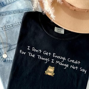 May include: Black t-shirt with the text "I Don't Get Enough Credit For The Things I Manage Not Say" and a cartoon frog graphic. The shirt is paired with blue jeans, a tan hat, and a silver necklace.