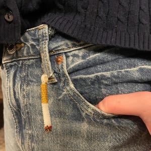 May include: A beaded cigarette charm in white, gold, and red hangs from the pocket of blue denim jeans. The charm is made of small beads and is attached to the jeans with a small loop.