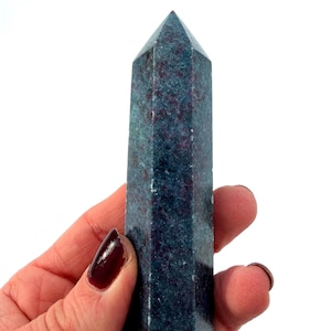 Puede incluir: Un obelisco de cristal azul oscuro con una parte superior puntiaguda. El cristal tiene un aspecto moteado con toques de rojo y se sostiene en una mano sobre un fondo blanco. El obelisco es una piedra preciosa y un objeto decorativo.