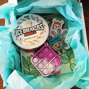 May include: A gift box containing a selection of items. There is a round container of Ice Breakers candy cane mints, a pink and white pop-it toy, a keychain with a smiling character and the text "You Rock", and stickers with positive affirmations.