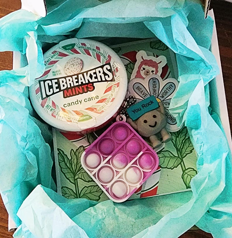May include: A gift box containing a selection of items. There is a round container of Ice Breakers candy cane mints, a pink and white pop-it toy, a keychain with a smiling character and the text "You Rock", and stickers with positive affirmations.