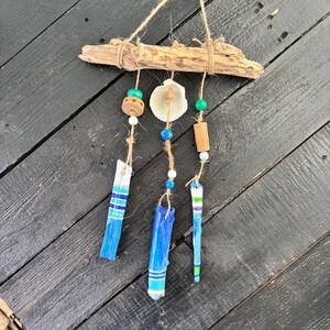 May include: A wall hanging featuring a piece of driftwood suspended by twine, with three dangling strands. The strands include painted blue and green wooden pieces, white and blue beads, a tan wooden bead, and a single white seashell.