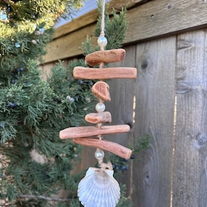 May include: A hanging ornament featuring five pieces of weathered brown driftwood and three white spherical beads strung together on a thin twine cord, finished with a single white scalloped seashell at the bottom of the vertical arrangement.