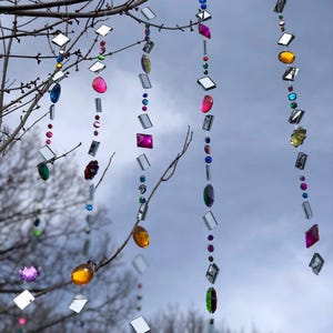Puede incluir: Adornos colgantes decorativos con gemas facetadas de colores y cuadrados espejados suspendidos de ramas de &aacute;rboles. Las gemas son de varias formas y colores. El fondo es un cielo nublado.
