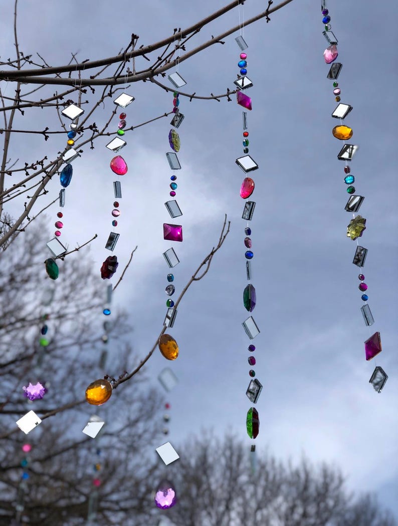 Puede incluir: Adornos colgantes decorativos con gemas facetadas de colores y cuadrados espejados suspendidos de ramas de &aacute;rboles. Las gemas son de varias formas y colores. El fondo es un cielo nublado.
