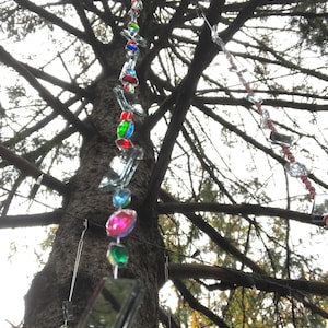 Puede incluir: Un adorno colgante decorativo para exteriores con cuentas facetadas de colores y peque&ntilde;os cuadrados reflectantes. El adorno est&aacute; suspendido de una rama de &aacute;rbol, con cuentas en tonos rojos, azules, verdes y rosas.