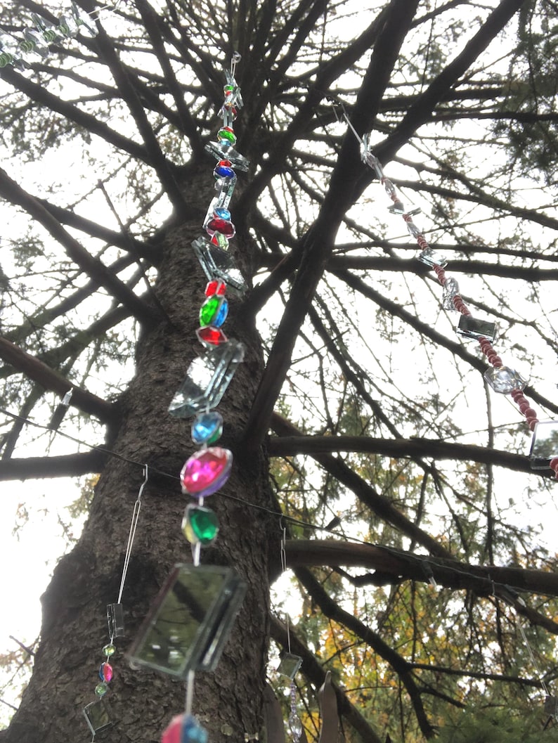 Puede incluir: Un adorno colgante decorativo para exteriores con cuentas facetadas de colores y peque&ntilde;os cuadrados reflectantes. El adorno est&aacute; suspendido de una rama de &aacute;rbol, con cuentas en tonos rojos, azules, verdes y rosas.
