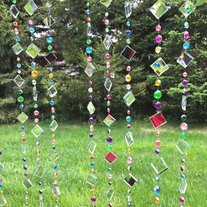 Puede incluir: Cortina decorativa de cuentas con piezas de vidrio transparentes en forma de diamante y cuadrado, intercaladas con cuentas redondas de colores. Las cuentas son en tonos de verde, rosa, morado, amarillo y azul. La cortina cuelga frente a un c&eacute;sped y &aacute;rboles verdes.