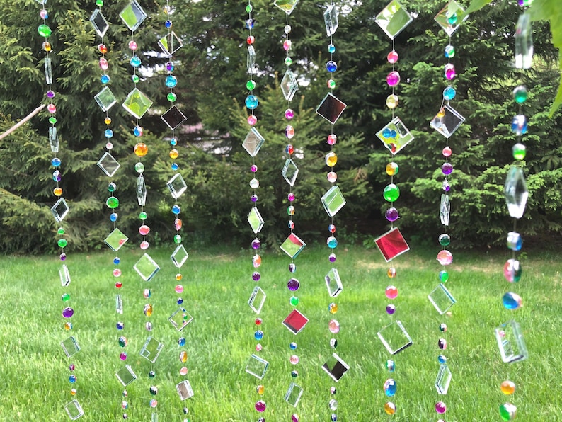 Puede incluir: Cortina decorativa de cuentas con piezas de vidrio transparentes en forma de diamante y cuadrado, intercaladas con cuentas redondas de colores. Las cuentas son en tonos de verde, rosa, morado, amarillo y azul. La cortina cuelga frente a un c&eacute;sped y &aacute;rboles verdes.