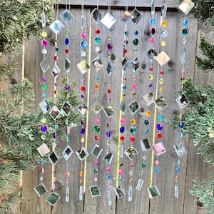 Puede incluir: Un carill&oacute;n decorativo con m&uacute;ltiples hilos de cuentas de colores y acentos de diamantes espejados. Las cuentas son de varios colores, incluyendo rojo, azul, verde y morado. El carill&oacute;n cuelga frente a una valla de madera.