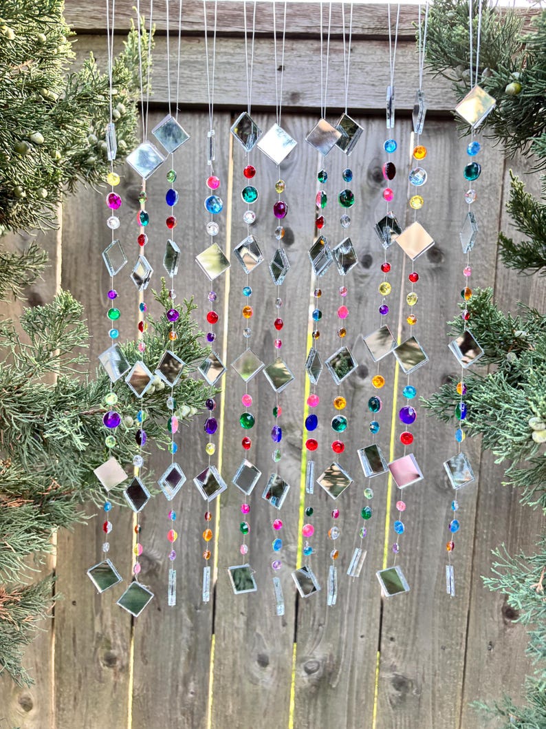 Puede incluir: Un carill&oacute;n decorativo con m&uacute;ltiples hilos de cuentas de colores y acentos de diamantes espejados. Las cuentas son de varios colores, incluyendo rojo, azul, verde y morado. El carill&oacute;n cuelga frente a una valla de madera.
