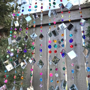 Puede incluir: Un carill&oacute;n decorativo con m&uacute;ltiples hilos de vidrio espejado cuadrado y transparente y cuentas de colores. Las cuentas son de varios colores, incluyendo rosa, azul, verde y morado. El carill&oacute;n cuelga frente a una valla de madera y ramas de hoja perenne.