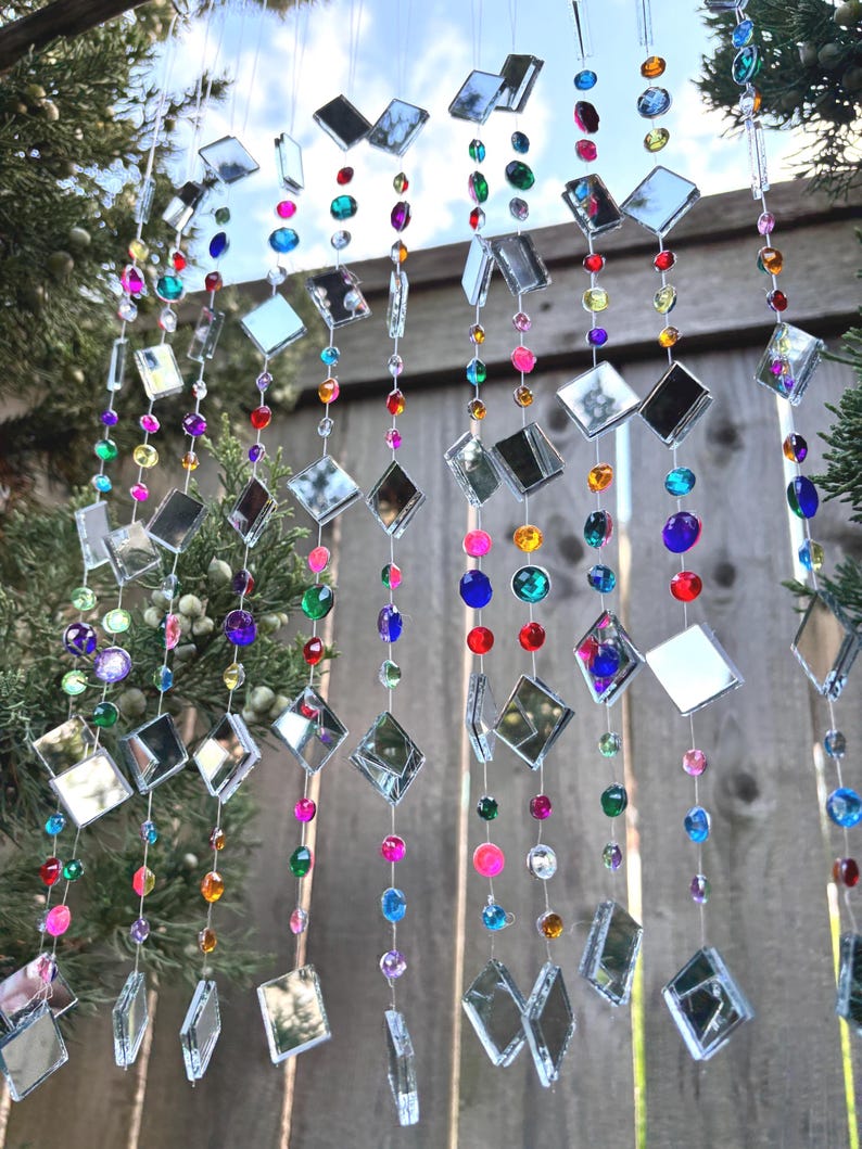Puede incluir: Un carill&oacute;n decorativo con m&uacute;ltiples hilos de vidrio espejado cuadrado y transparente y cuentas de colores. Las cuentas son de varios colores, incluyendo rosa, azul, verde y morado. El carill&oacute;n cuelga frente a una valla de madera y ramas de hoja perenne.