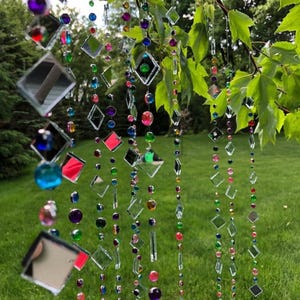 Puede incluir: Adorno colgante decorativo para exteriores con hilos de cuadrados de vidrio transparente y reflectante y cuentas de colores. Las cuentas son en tonos rosa, morado, verde, azul y amarillo. El adorno cuelga de una rama de &aacute;rbol.