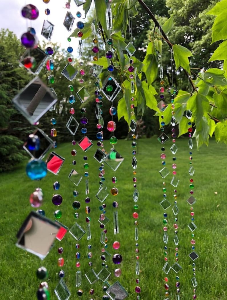 Puede incluir: Adorno colgante decorativo para exteriores con hilos de cuadrados de vidrio transparente y reflectante y cuentas de colores. Las cuentas son en tonos rosa, morado, verde, azul y amarillo. El adorno cuelga de una rama de &aacute;rbol.