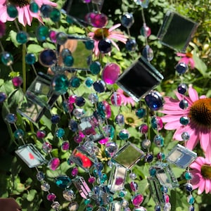 Puede incluir: Una guirnalda decorativa con baldosas de espejo cuadradas y cuentas de colores en tonos de azul, verde y rosa. La guirnalda se muestra sobre un fondo de flores rosas y follaje verde.
