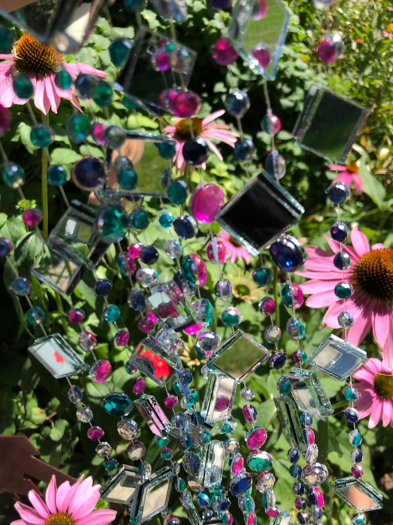 Puede incluir: Una guirnalda decorativa con baldosas de espejo cuadradas y cuentas de colores en tonos de azul, verde y rosa. La guirnalda se muestra sobre un fondo de flores rosas y follaje verde.