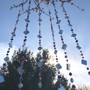 Puede incluir: Cadenas decorativas colgantes de vidrio con cuentas rojas, azules y plateadas y piezas de vidrio en forma de diamante. Las cadenas est&aacute;n suspendidas de ramas de &aacute;rboles contra un cielo azul, creando una decoraci&oacute;n exterior caprichosa.