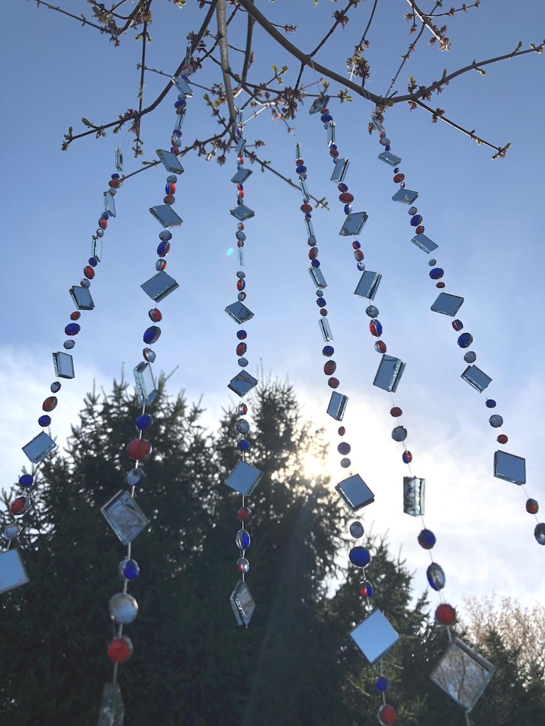Puede incluir: Cadenas decorativas colgantes de vidrio con cuentas rojas, azules y plateadas y piezas de vidrio en forma de diamante. Las cadenas est&aacute;n suspendidas de ramas de &aacute;rboles contra un cielo azul, creando una decoraci&oacute;n exterior caprichosa.