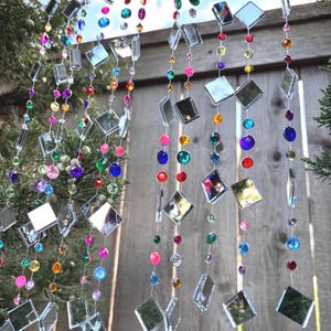 Puede incluir: Cadenas decorativas colgantes con cuadrados espejados y cuentas coloridas en tonos rosa, azul, verde, morado y naranja. Las cadenas cuelgan frente a una valla de madera y vegetaci&oacute;n.